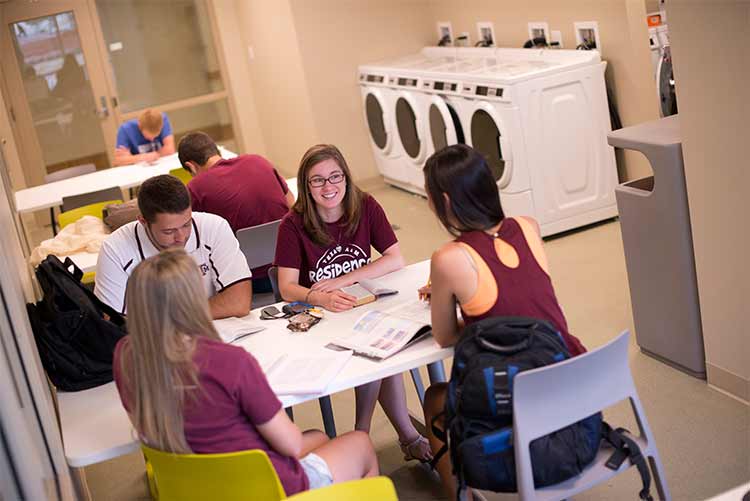 Picture of six teens studying at a table in the laundry room. Four dryers are pictured in the image. 