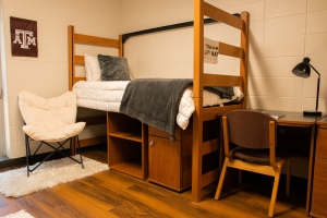 Commons Hall lofted twin bed, with storage unit underneath, next to a desk and chair. Next to the lofted bed is a White decorative chair on top of a White rug. On the desk is a Black lamp.