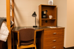Commons Hall desk and chair, next to a dresser with a bookcase on top. On the desk is a black lamp. The book case has a black coffee maker, and a white Texas A&M coffee cup  on the top shelf. A framed photo sits on top of the book case.