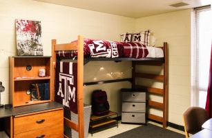 Interior of a decorated Dunn dorm. A lofted bed with A&M bedding is shown, with a shoe rack, laundry basket, and storage drawers underneath.  Next to the lofted bed is a desk, with a bookshelf on top.