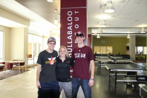 Three students standing with their arms linked, smiling at the camera,  in front of the "Hullabaloo Hangout" sign in the Hullabaloo game room.