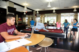 A student studying at their laptop, while other students play pool in the Hullabaloo game room