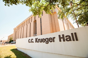 The C.C Krueger Hall Sign with the outside of Krueger Hall in the background, surrounded by trees.