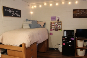 Decorated Krueger Hall dorm room.  Decorative lights hang above the lofted twin bed with many pillows. Above the bed is a decorative poster that says "in everything give thanks,". Next to the bed  are photos pinned to a photo board, and a microwave and fridge.