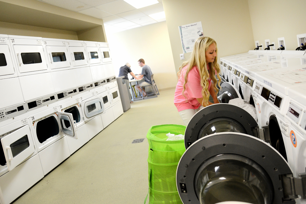 A female resident doing her laundry in the laundry room of a residence hall. Two students study at a table in the background