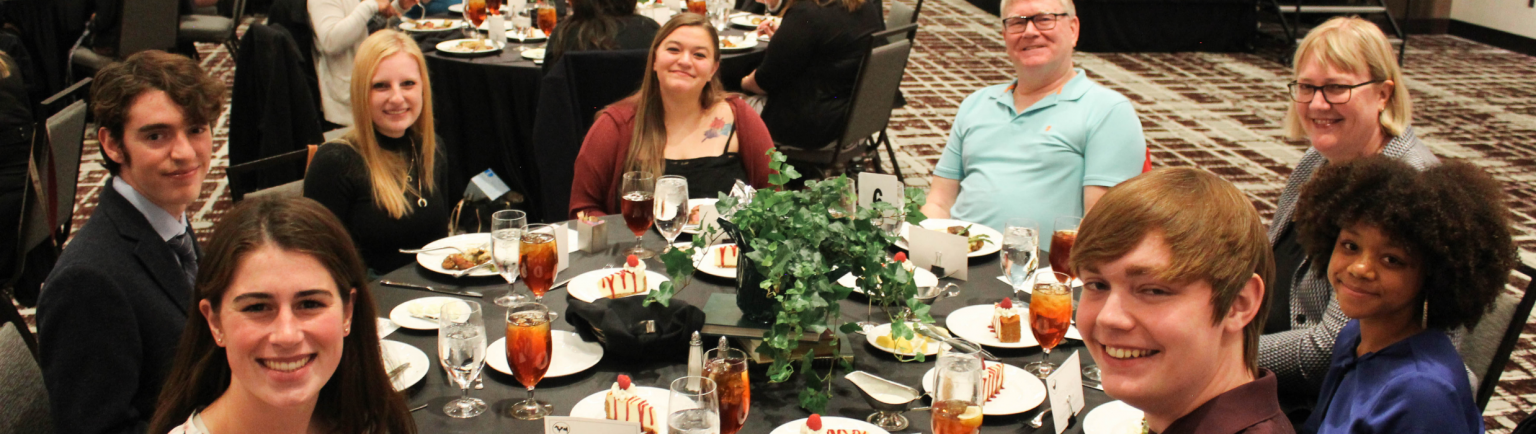 students and faculty sitting together at a table at an awards ceremony, smiling at the camera