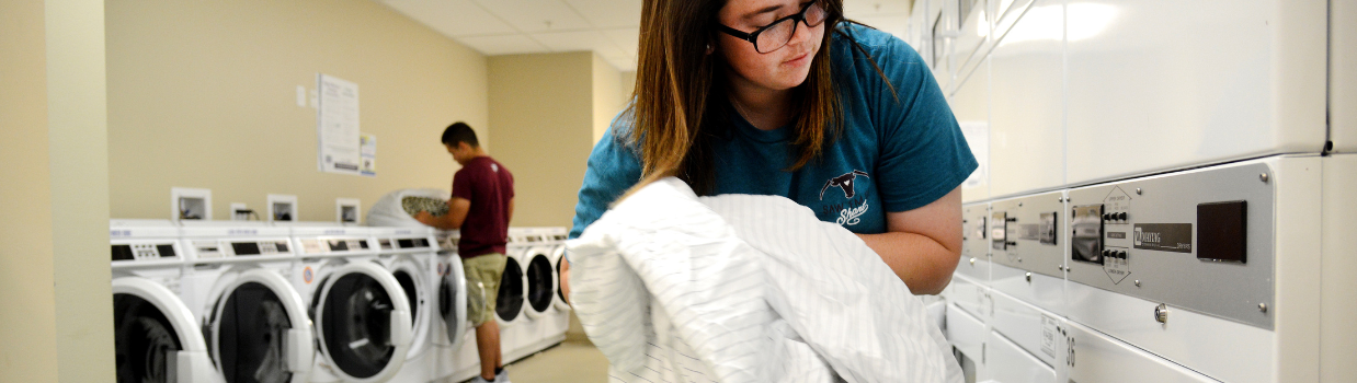 Close up of student putting her white sheets into a laundry machine in a residence hall laundry room. In the background is another student who is also doing their laundry.